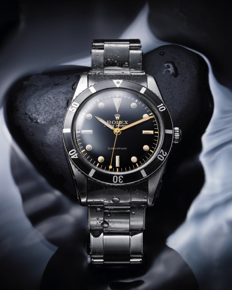 Vintage Rolex Submariner, black dial and bezel. The watch is lying against a black rock with water on it.