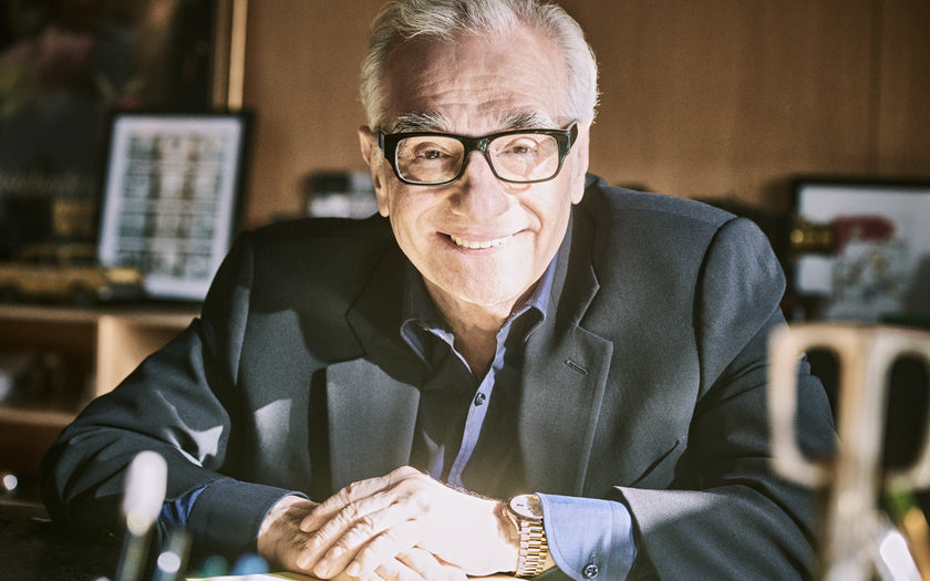 A distinguished gentleman with black thick framed glasses. He is wearing a suit and blue shirt, as well as a Rolex Day-Date.