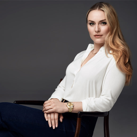 Rolex testimonee Lindsey Vonn sitting in a chair in a white blouse. She is wearing a Rolex Day-Date in yellow gold with a diamond bezel.