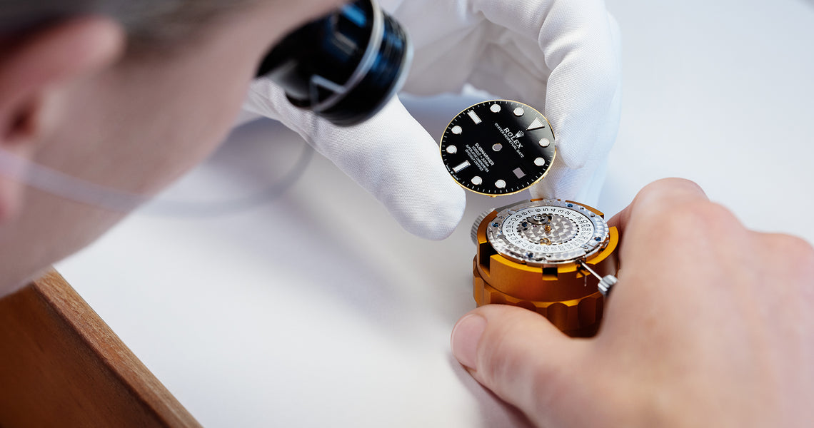 A Rolex watch technician wearing a white glove and wearing a loupe affixing a black dial to a Rolex movment.