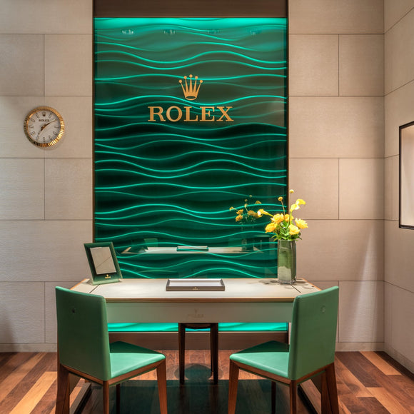 A large green Rolex background wall piece with a selling desk and 2 chairs.